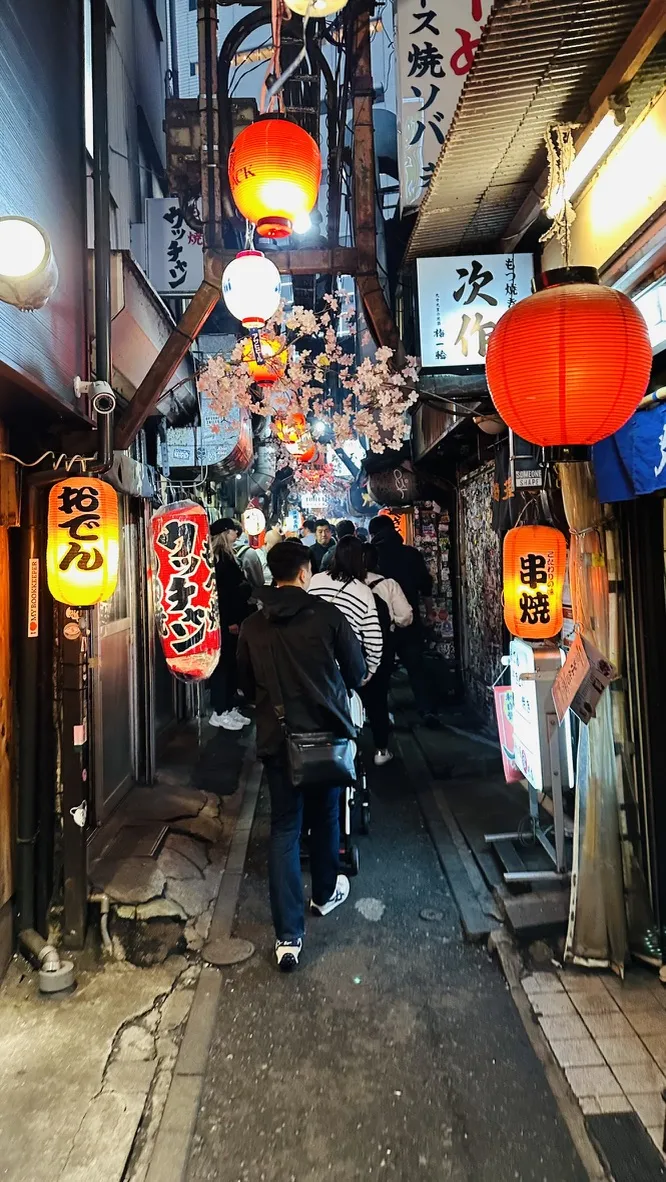 A narrow alleyway bustling with people, illuminated by vibrant red lanterns and adorned with cherry blossom decorations. Various restaurant signs in Japanese line the walls, creating a lively and inviting atmosphere.