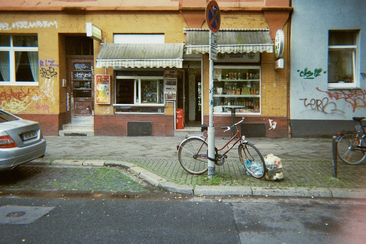A vintage bicycle leans against a street pole in front of a colorful building with a small storefront. The walls are adorned with graffiti, and nearby, a parked car is visible along the cobblestone road.