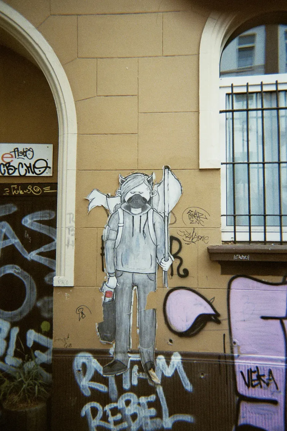 A large, monochrome graffiti mural depicts a person with a long beard and a backpack, holding a flag in one hand and a drink in the other. The mural is set against a textured, tan wall covered in various colorful tags and graffiti.