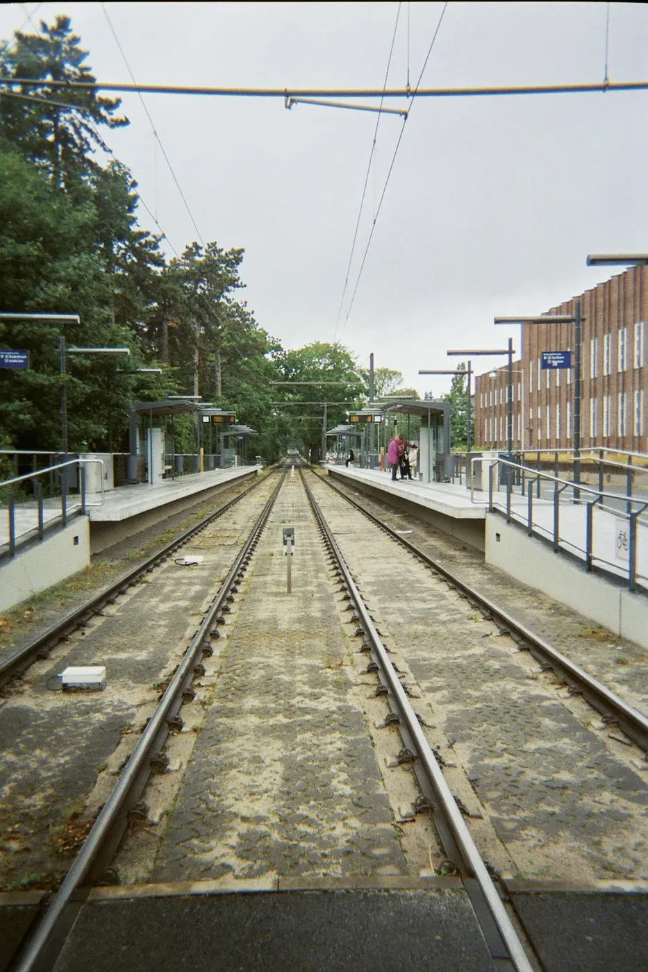 Two parallel railway tracks run through a station with platforms on either side, surrounded by trees and a modern building. The sky is overcast, and a person is visible on one of the platforms.