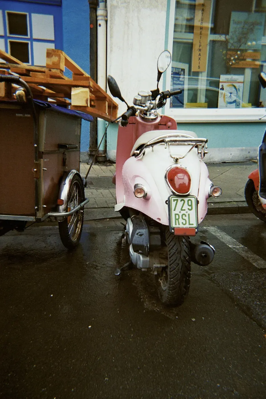 A pink scooter parked next to a small cart with wooden planks on top. The background features a colorful building with a blue facade. The ground is wet, suggesting recent rain.