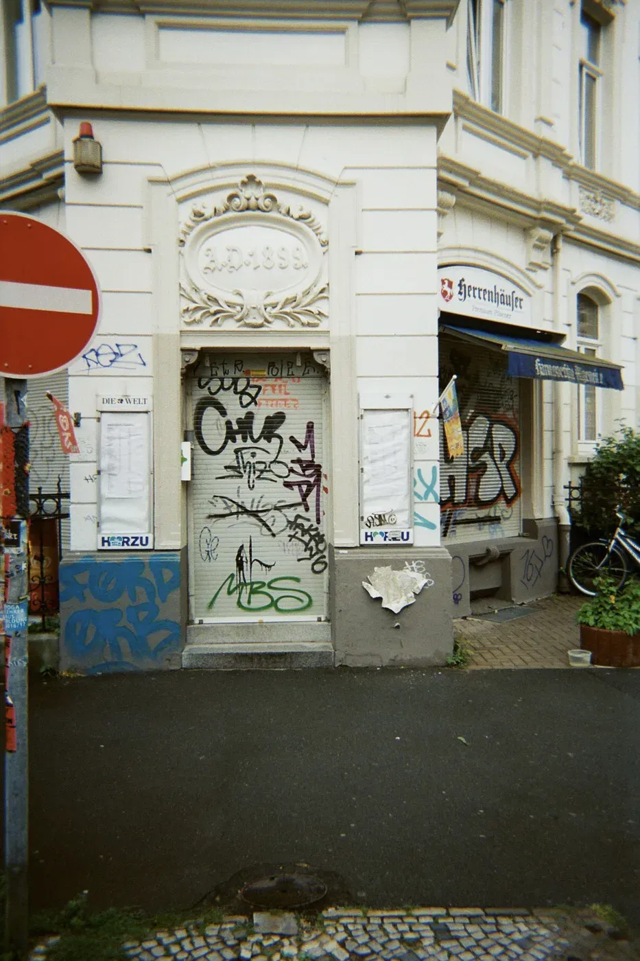A partially obscured entrance with ornate architrave features a mix of graffiti and posters on the walls. A nearby sign indicates a no entry direction, while a bicycle leans against the building, and potted plants add a touch of greenery to the scene.