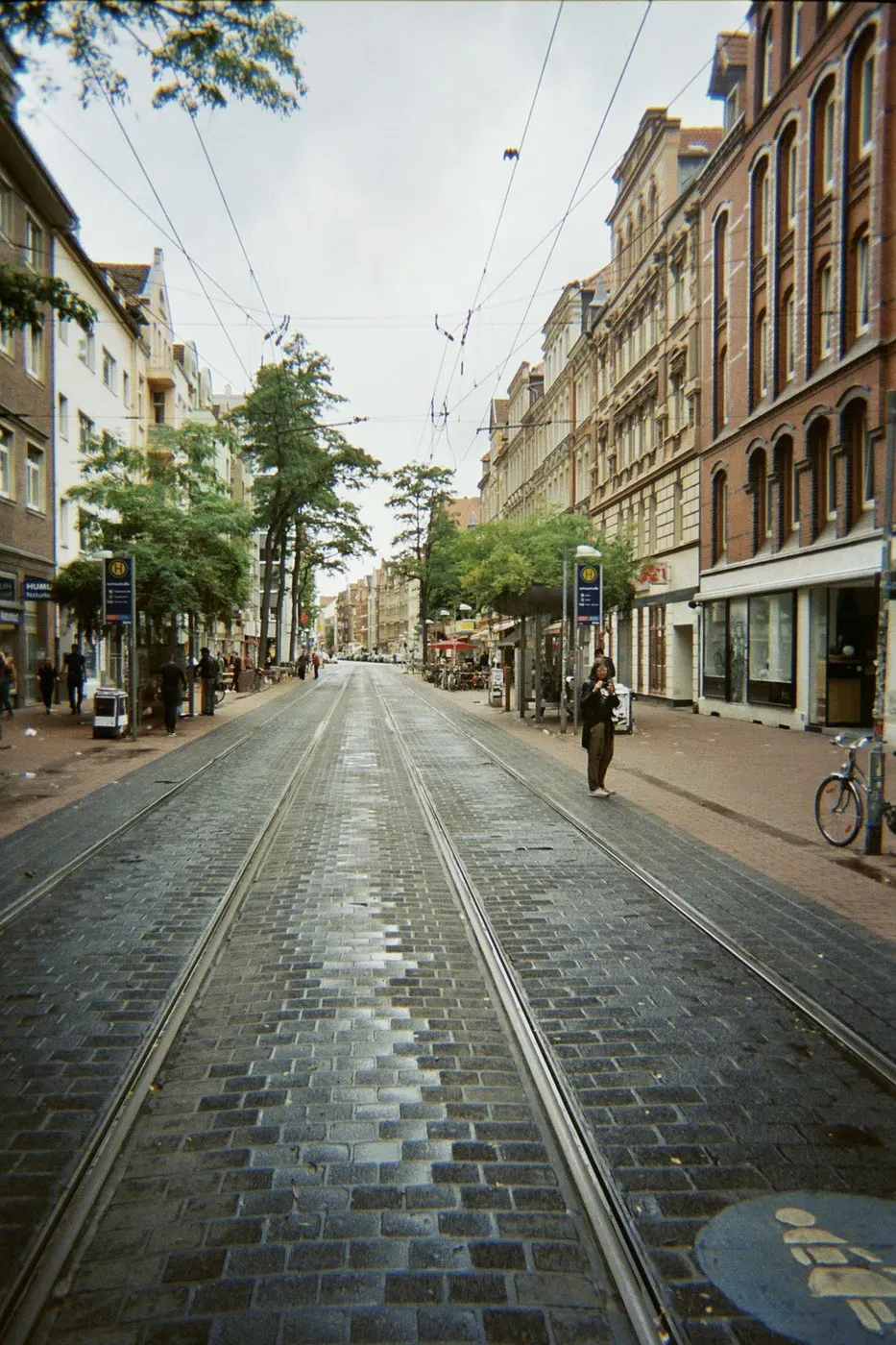 A wide cobblestone street runs through a city, lined with trees and historic buildings on either side. Tram tracks can be seen down the center of the road, while a few pedestrians and bicycles are visible along the sidewalk.
