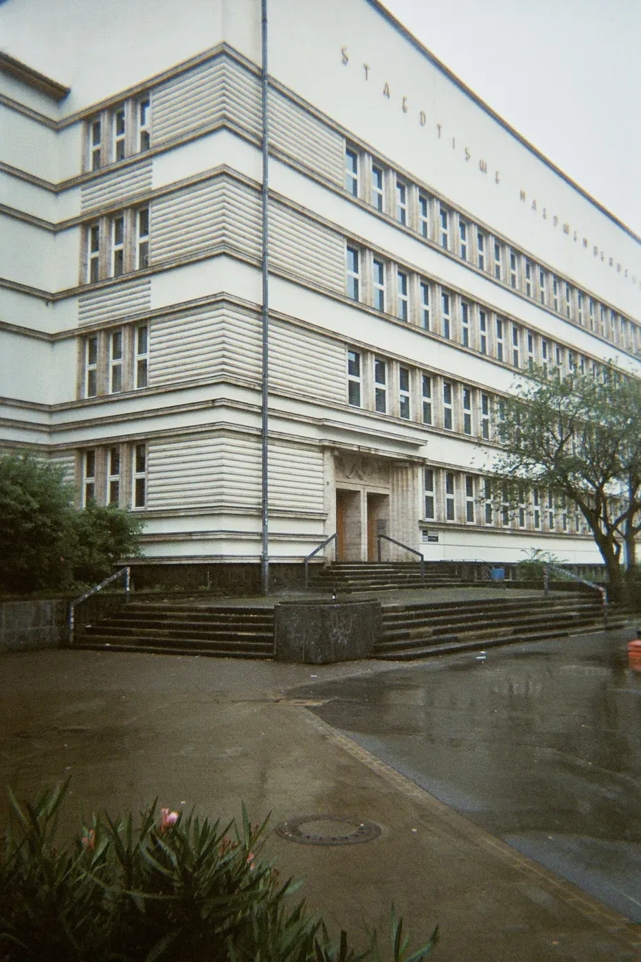 A large, modernist building with a textured facade features multiple rectangular windows. The entrance has a set of stairs leading up to it, and the scene shows a damp, overcast day with a few trees and greenery in the foreground.