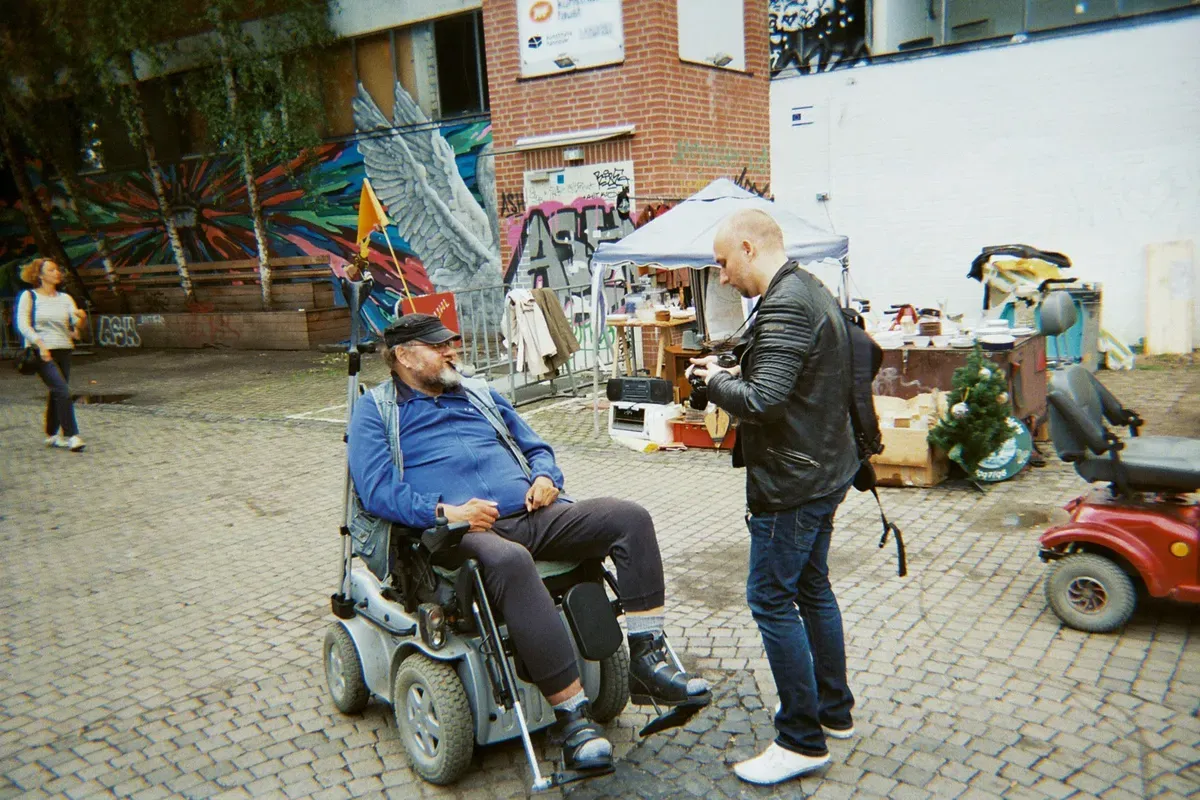 A man in a wheelchair is seated on a cobblestone street, engaging with another man who is holding a camera and taking his picture. In the background, there are colorful murals and various outdoor market stalls.