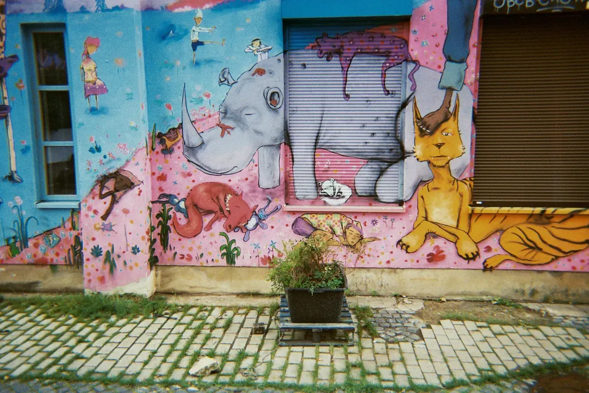 A vibrant mural covers a building wall, depicting whimsical animals including an elephant, a fox, and a cat, surrounded by colorful patterns and playful illustrations. A small plant in a pot sits on a cobblestone path in front of the mural.