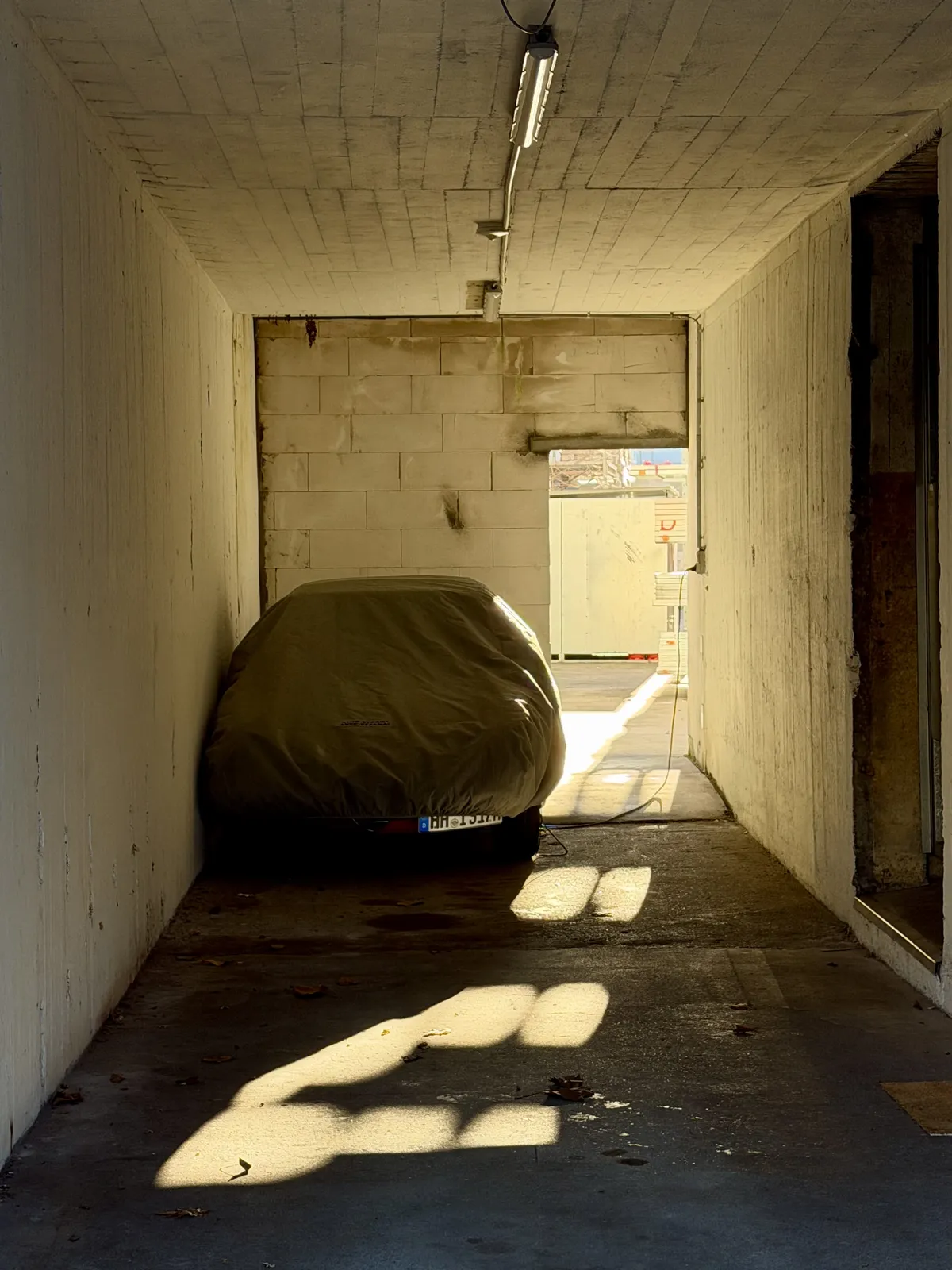 A covered car is parked in a narrow, well-lit passageway with white walls. The sunlight creates shadows on the ground, highlighting the contours of the car and the texture of the wall.