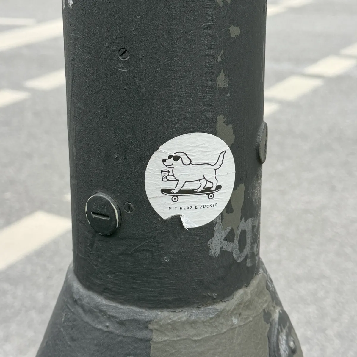 A sticker on a gray lamppost depicts a cartoon dog riding a skateboard while holding a drink. The design is simple with bold black lines on a white background.