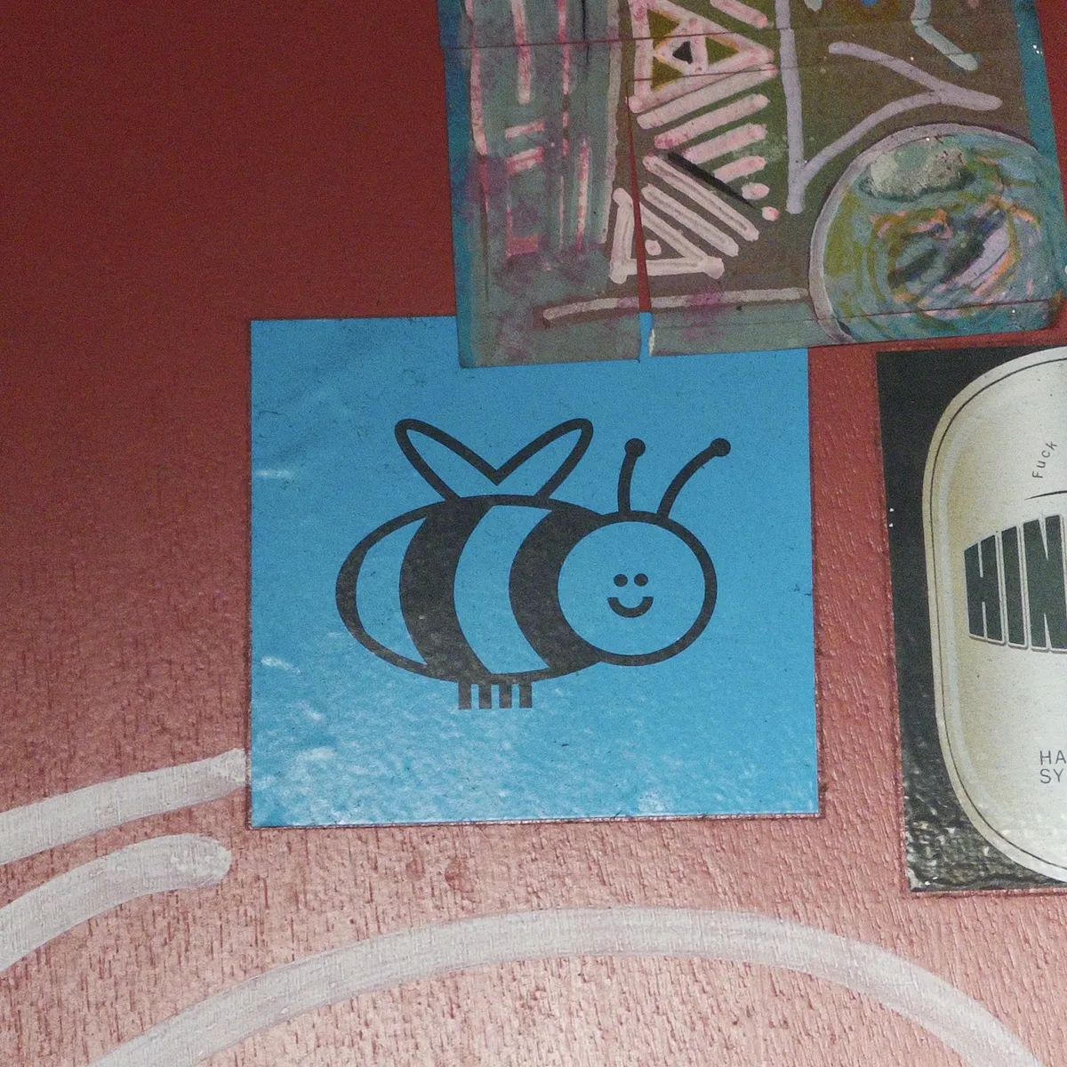 A blue square with a cartoon-style bee illustration is displayed on a red wall. Surrounding it are abstract art pieces, including colorful shapes and lines.