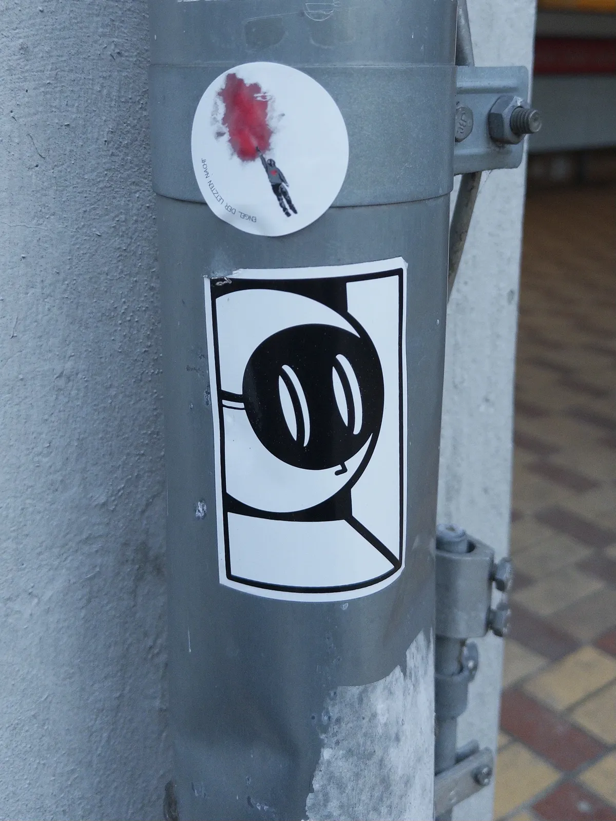A grey metal pole features a black and white sticker depicting a cartoonish character with large eyes and a simple face, positioned in a square outline. A small circular sticker with a red splash is also visible above the main sticker.