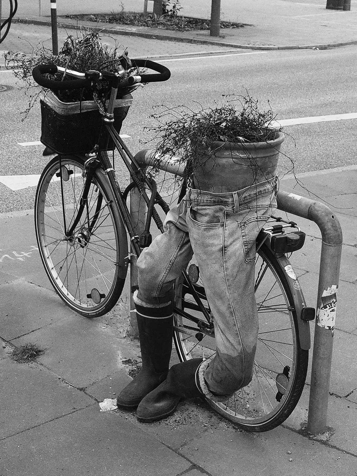 A bicycle is creatively transformed with potted plants in place of its handlebars and a pair of jeans and rubber boots positioned on its frame, giving the appearance of a person. The scene is captured in black and white.