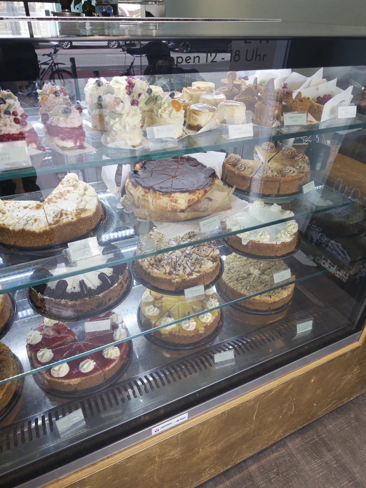 A glass display case filled with various types of cakes and cheesecakes, including fruity toppings, chocolate ganache, and decorative frosting. The cakes are arranged on multiple shelves, showcasing a colorful assortment of flavors and styles.
