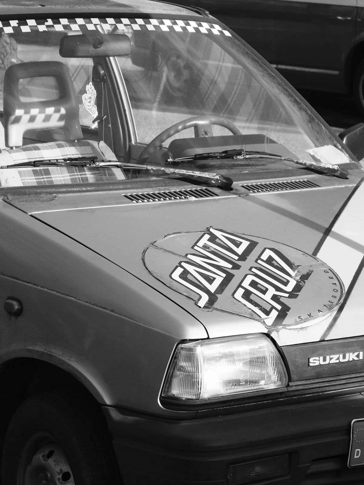 A close-up of a red Suzuki car with a large circular sticker on the hood that says 'SANTA CRUZ.' The interior features plaid upholstery, visible through the windshield. The image is in black and white.