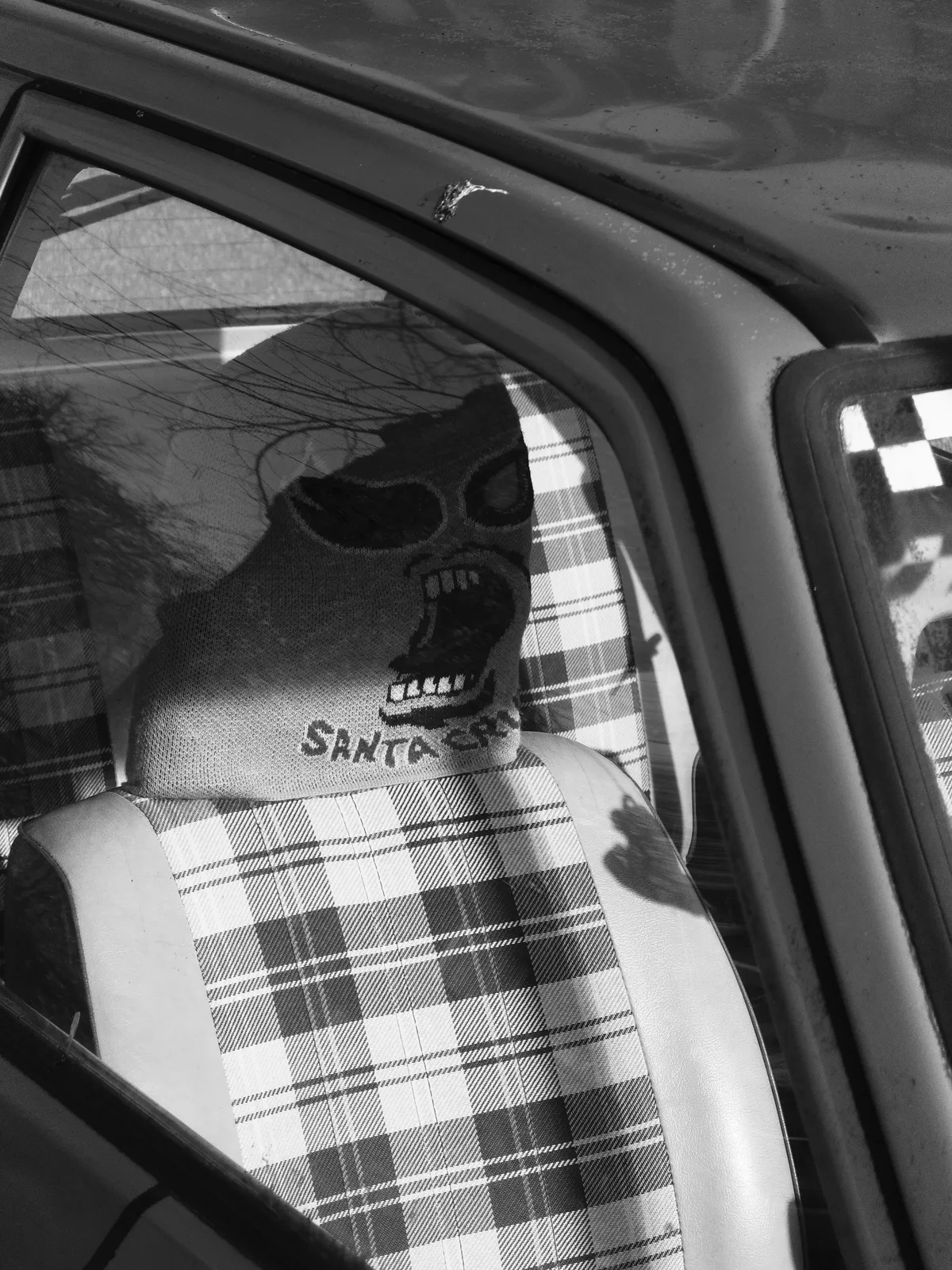 A car seat covered with a plaid fabric shows a skull design embroidered on a seat cover. The interior is dimly lit, highlighting the patterns of the seat and the shadowy effect from outside.