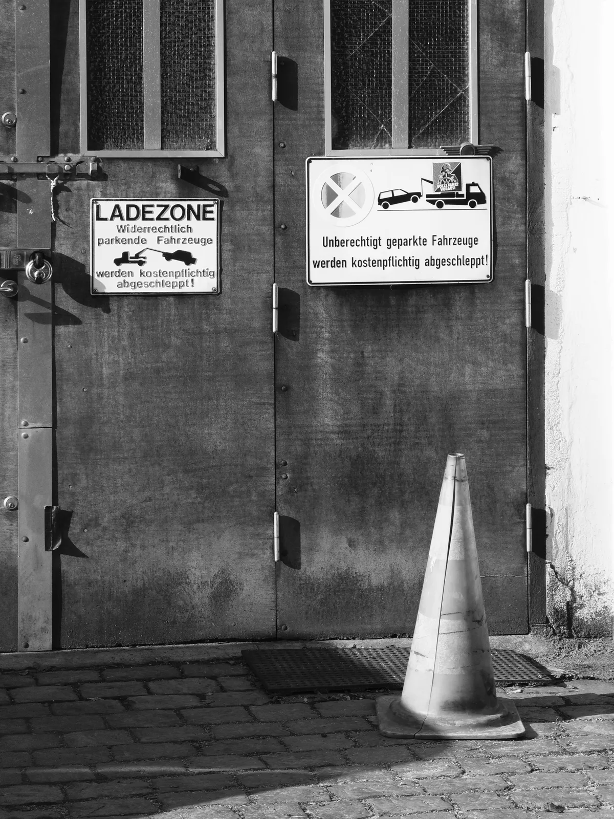 A metal door with two signs attached: one indicating a loading zone and the other warning that improperly parked vehicles will be towed, along with a traffic cone placed in front of the door. The image is in black and white, highlighting the textures of the door and signs.