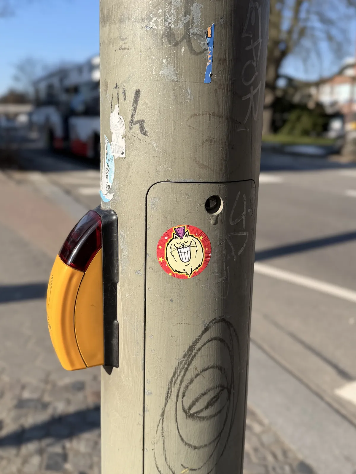 A weathered utility pole is shown with a bright yellow pedestrian crossing button on the left. The pole features various graffiti and stickers, including a prominent sticker of a grinning cartoon character with a white face and spiky hair.