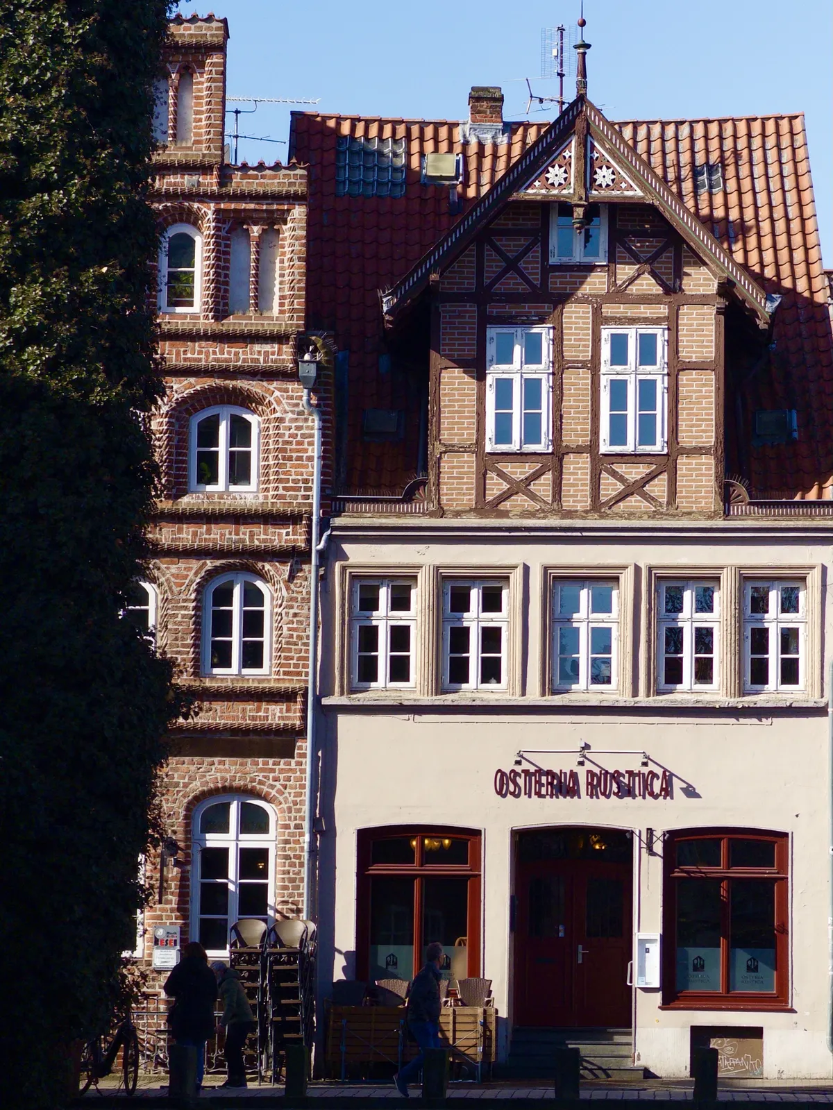 A historic building with a mix of architectural styles, featuring half-timbered elements and a modern facade. People are seen walking by.