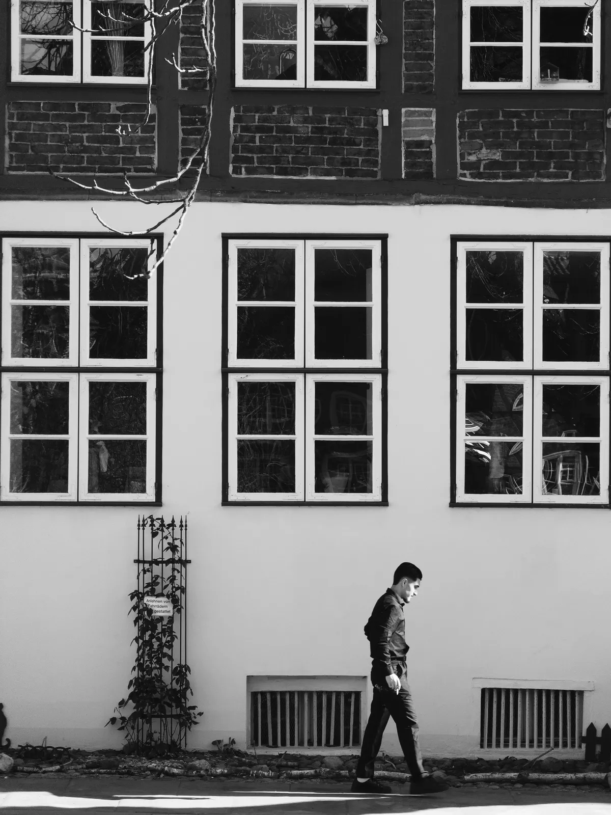 A person walks along a sidewalk in front of a building with large, rectangular windows. The scene is captured in black and white, highlighting the contrast between the individual and the architecture of the structure.
