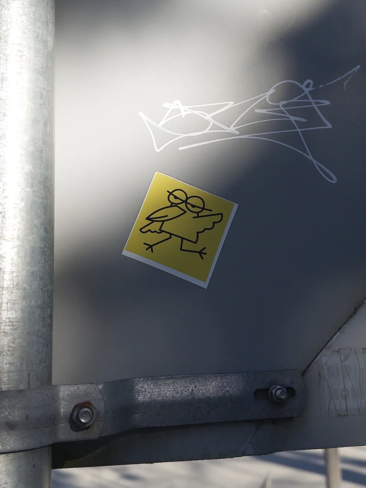 A yellow sticker featuring a cartoonish bird design with large glasses sits on a gray metal surface, accompanied by some white graffiti nearby. The bird has a playful expression with its wings slightly outstretched.