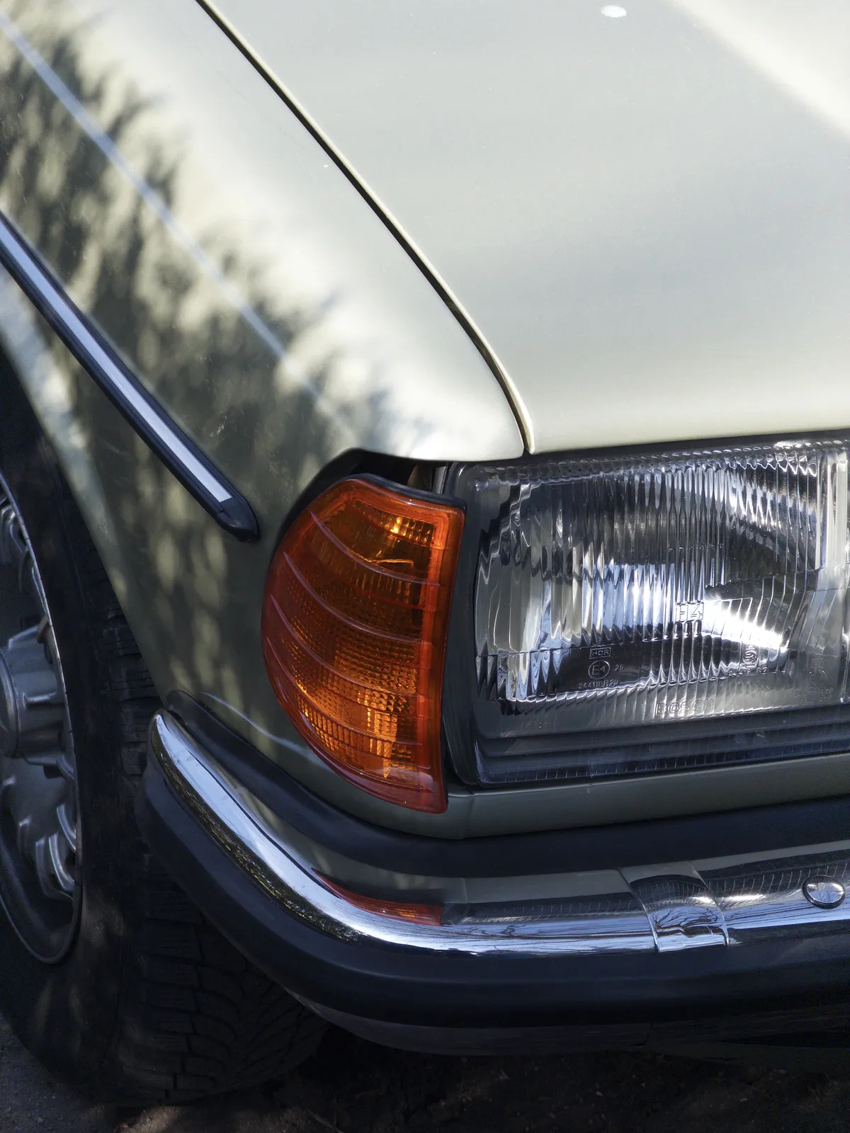 Close-up of a vintage car's front corner, featuring a round headlight and an orange turn signal indicator. The car's paint is a soft green, with a shiny chrome trim along the fender.