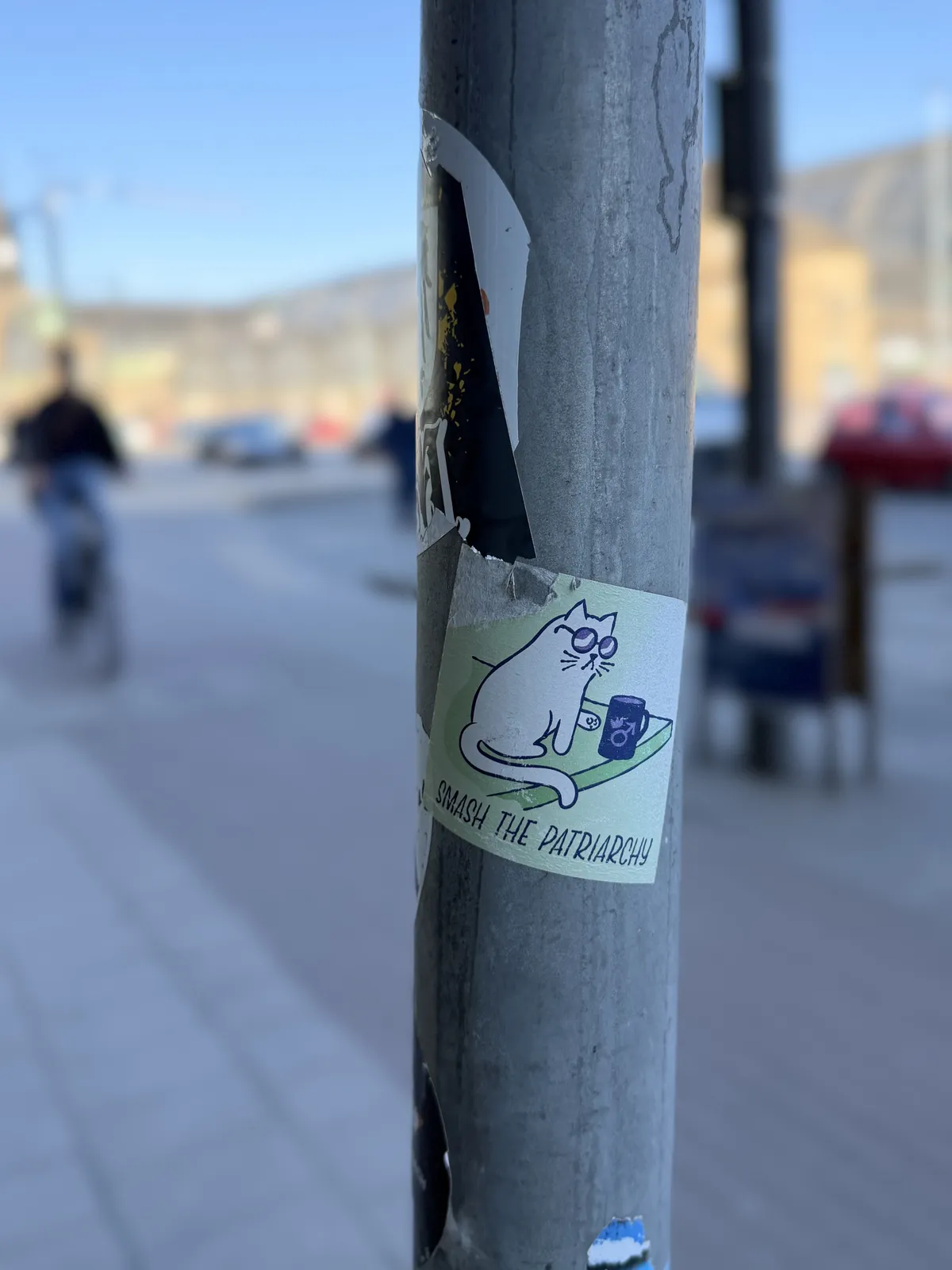 A metal pole in an urban setting features a sticker depicting a cartoon cat sitting at a table with a drink, accompanied by the text 