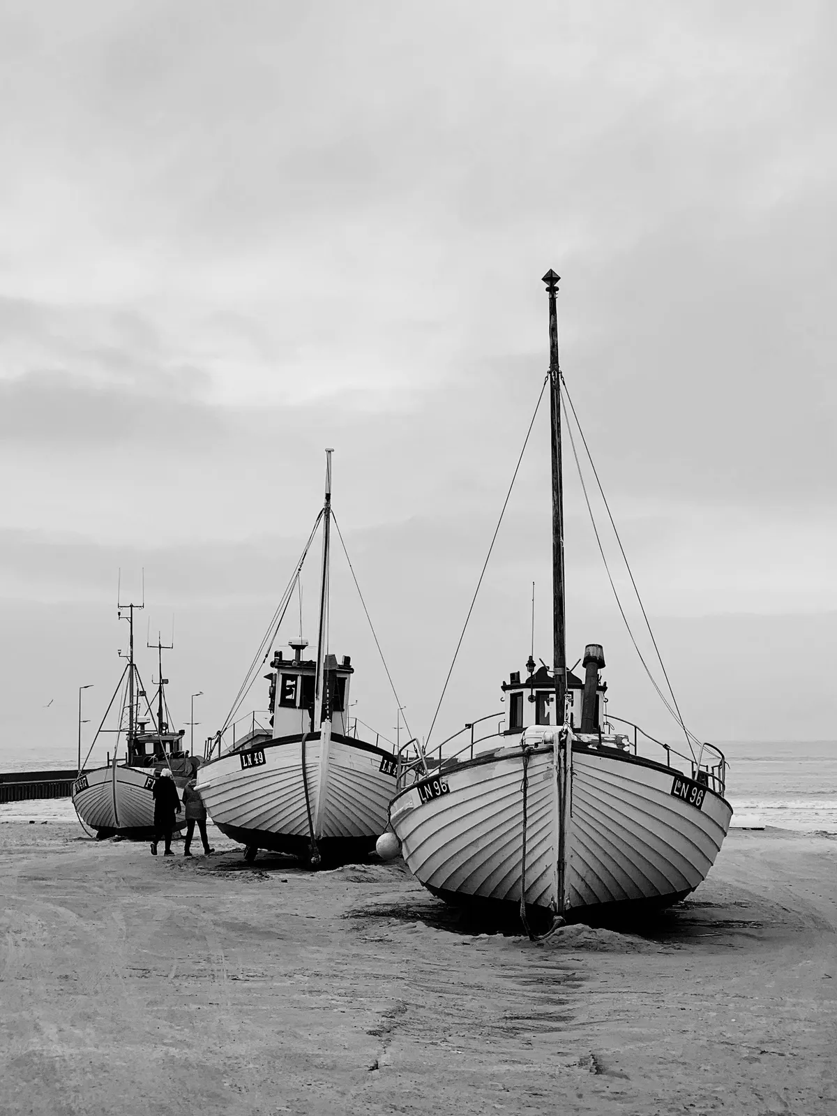Three fishing boats are anchored on a sandy beach, with two figures walking between them. The scene is presented in black and white, with a cloudy sky in the background.