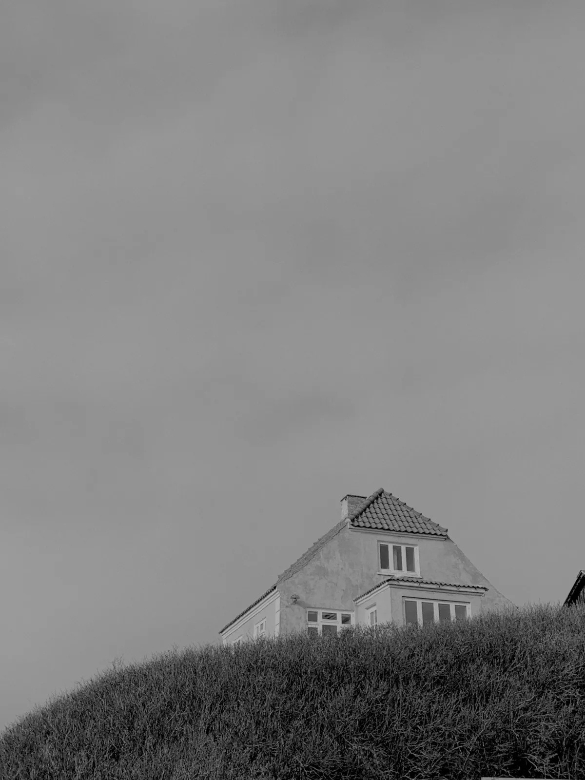 A house with a sloped roof is perched on top of a grassy hill, set against a cloudy sky. The image is in black and white, emphasizing the contrast between the building and the landscape.