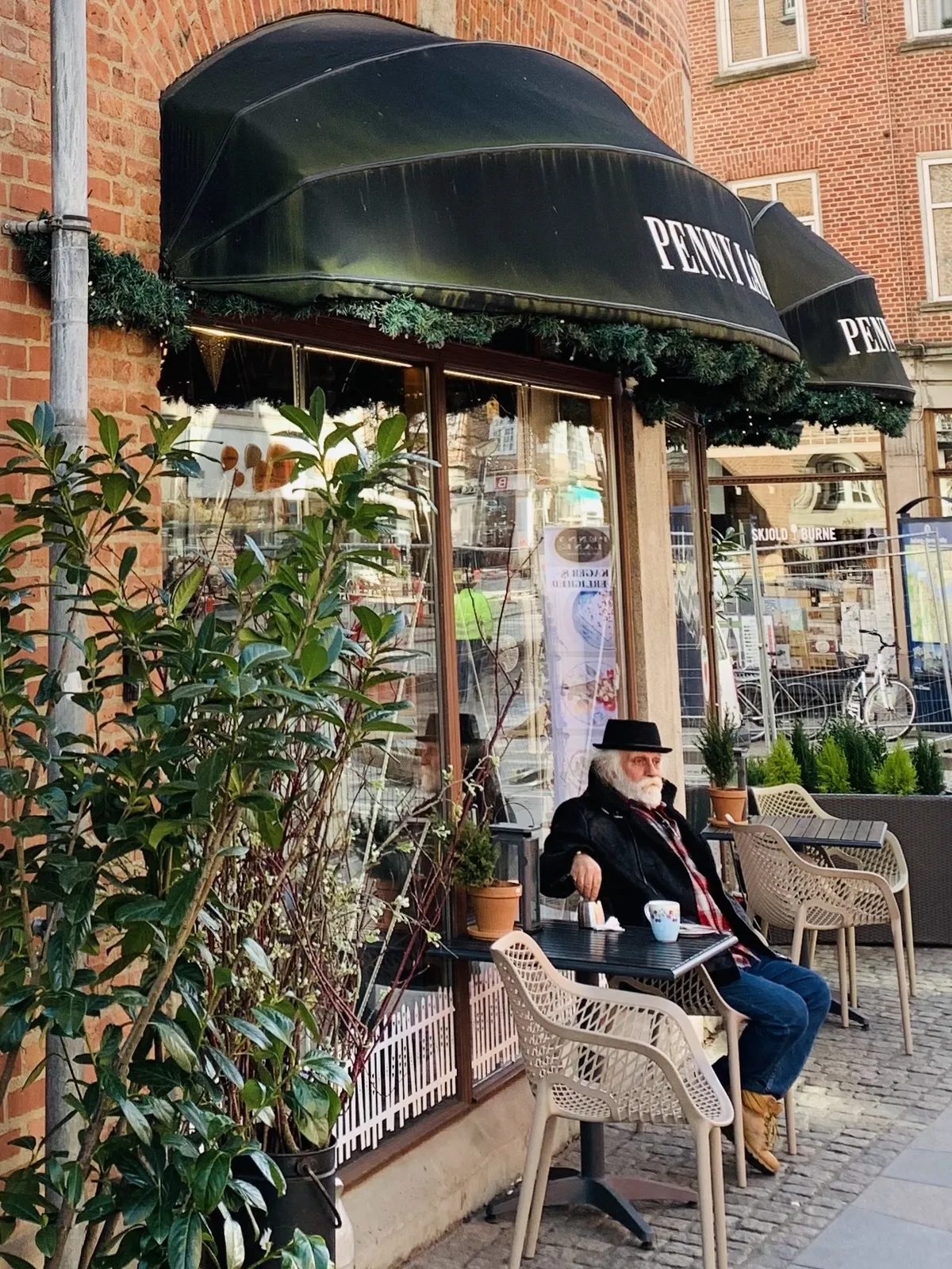 A man in a hat sits at a small outdoor café table in front of a brick building, with plants and decorative elements around the entrance. The café awning is dark, and the scene includes a cobblestone street and a glimpse of activity in the background.
