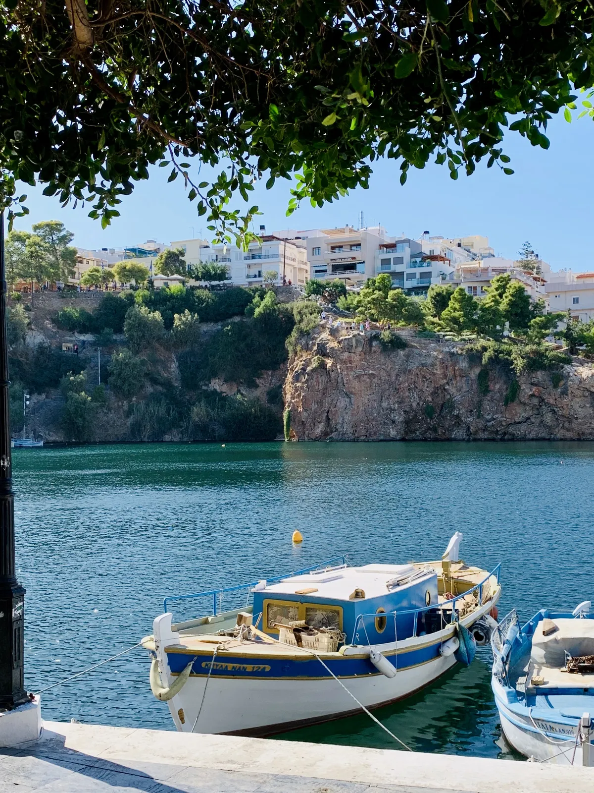 A serene harbor scene featuring two boats moored in calm waters, with a rocky cliff and green trees in the background. Colorful buildings line the top of the cliff under a clear blue sky.