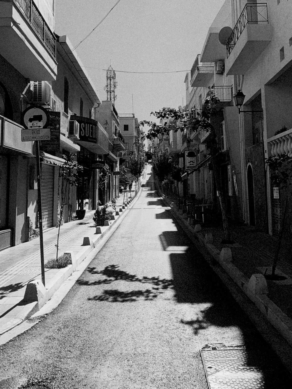 A narrow, empty street lined with shops and buildings, casting long shadows on the pavement. The scene is in black and white, highlighting the contrast between light and shadow, with a slight incline leading up the street.