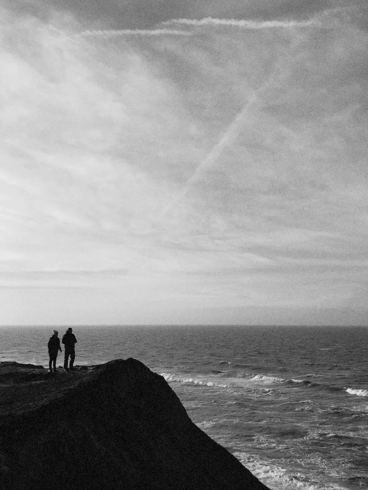 Two silhouettes of people walk along a cliff edge, overlooking a calm ocean with gentle waves. The sky is filled with soft clouds, and crisscross vapor trails can be seen above.