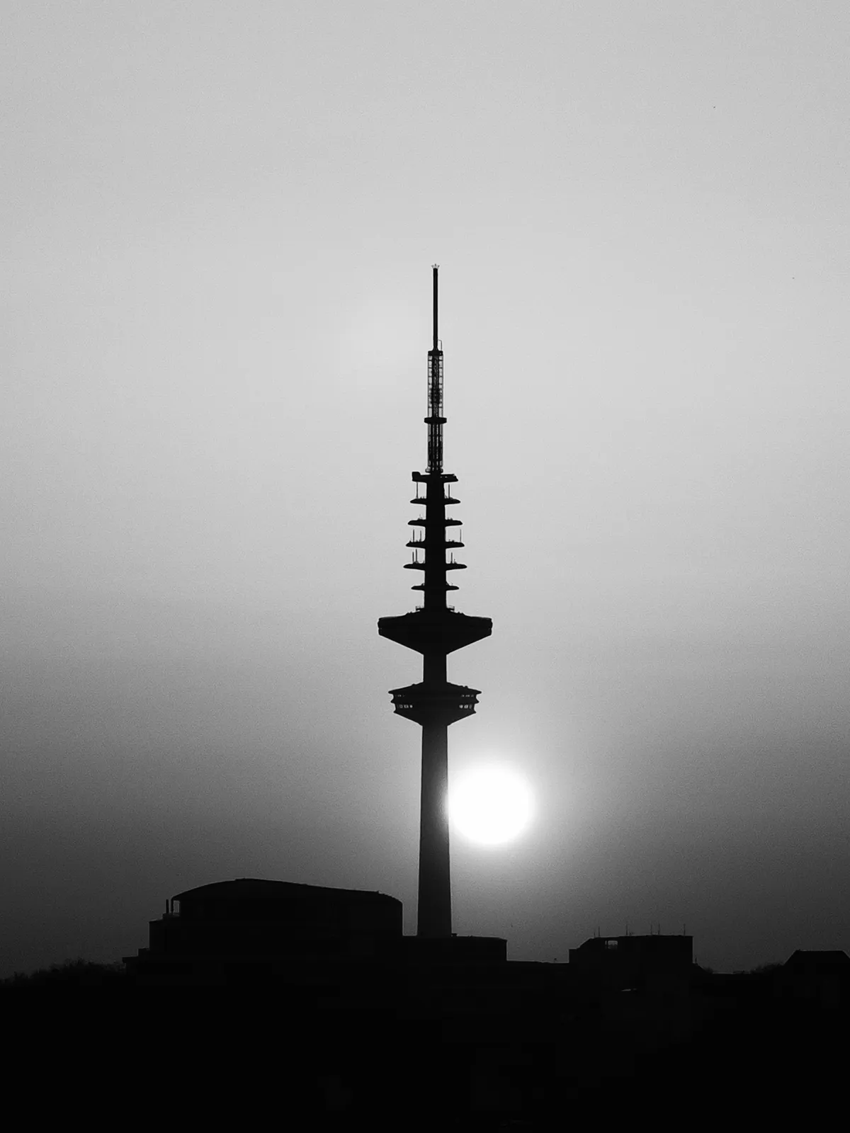 A silhouette of a tall communication tower against a hazy sky, with the sun partially obscured behind it. The scene captures a tranquil sunset atmosphere.