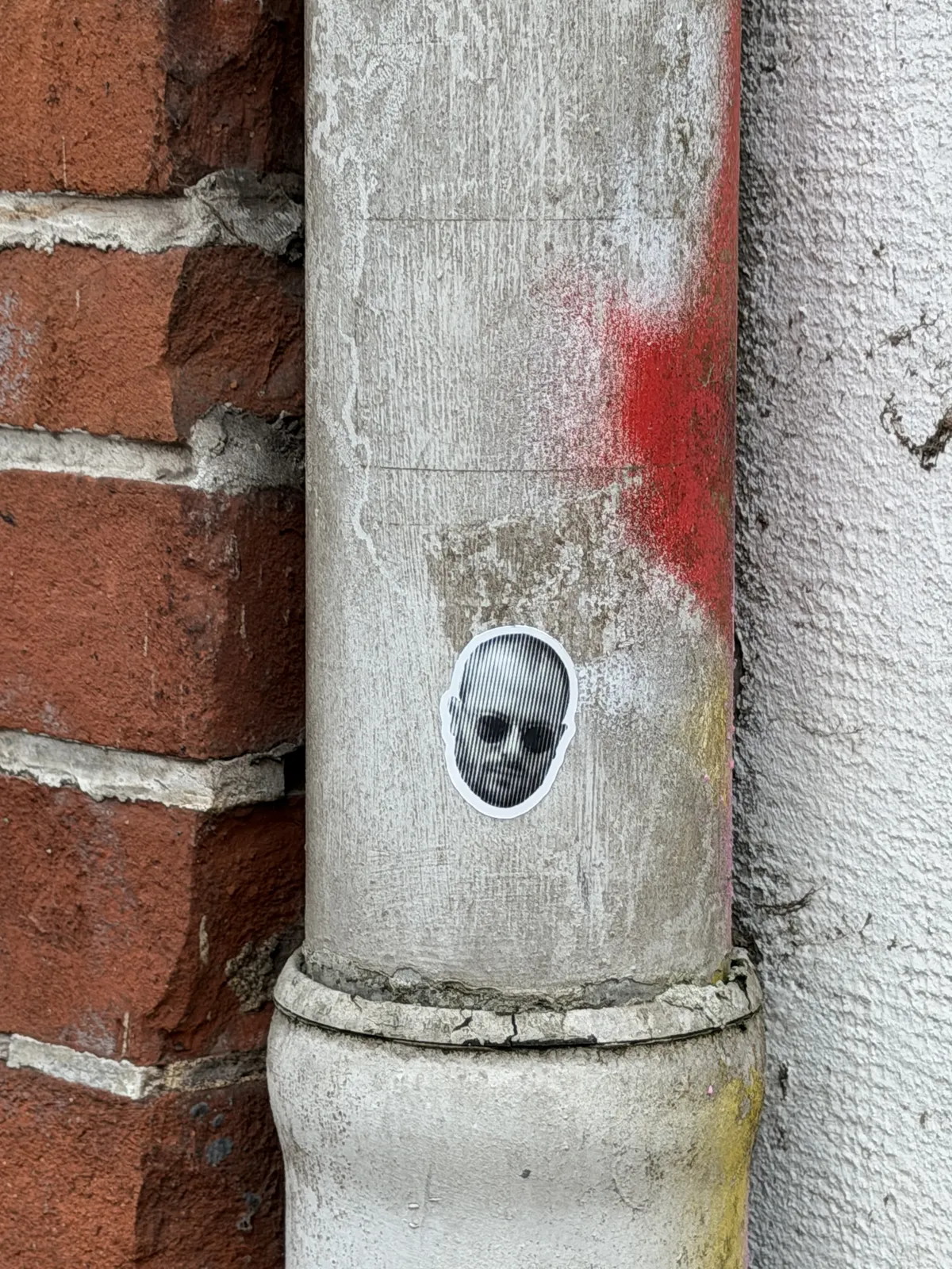 A small black and white sticker featuring a distorted face design is affixed to a gray, weathered metal pipe. The background includes red graffiti and a brick wall.