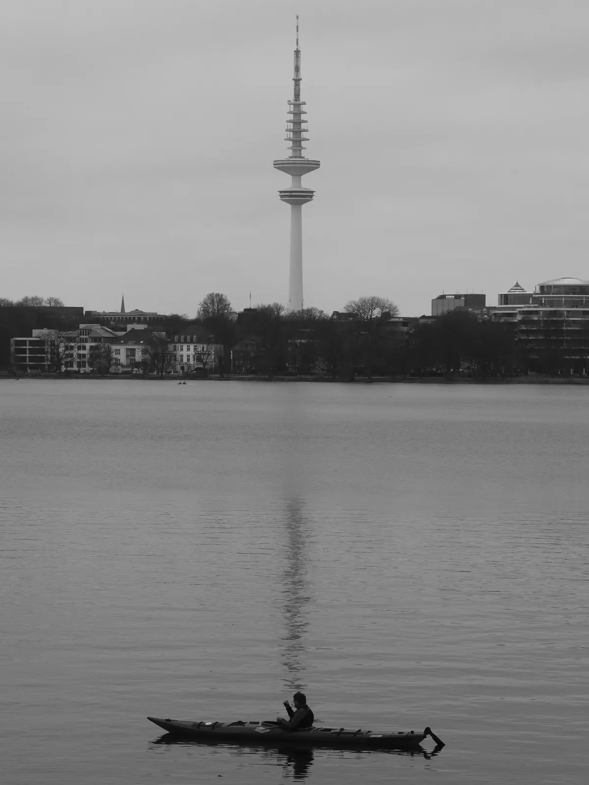 A person sits in a small boat on a calm, reflective body of water, with a tall television tower in the background. The scene is presented in black and white, capturing a serene atmosphere.