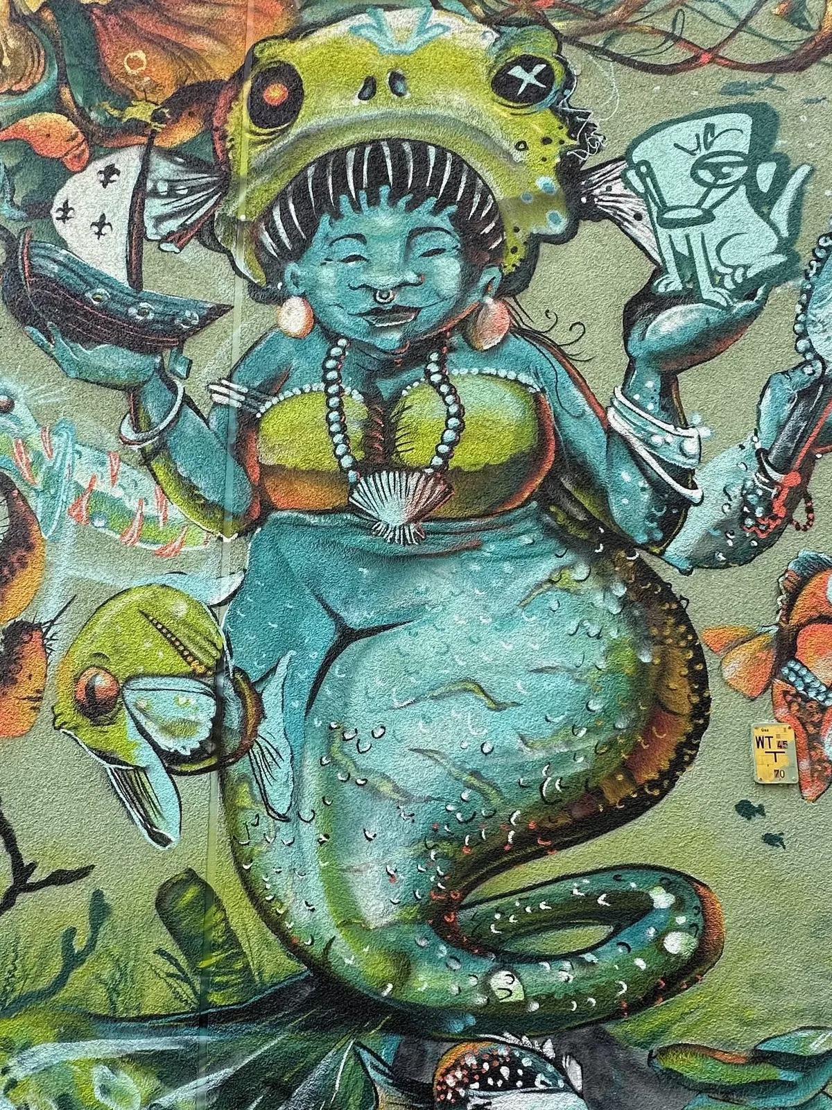 A colorful mural depicting a mermajd-like figure with a fish head and a vibrant green tail. The character holds a cup in one hand and is adorned with jewelry, surrounded by aquatic motifs and detailed marine life in the background.
