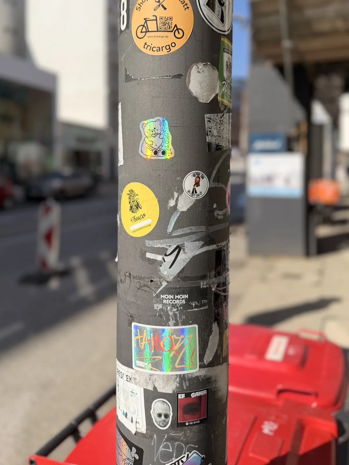 A gray utility pole covered in a variety of colorful stickers, including logos and illustrations. The background shows a city street with buildings and a red cart in the foreground.