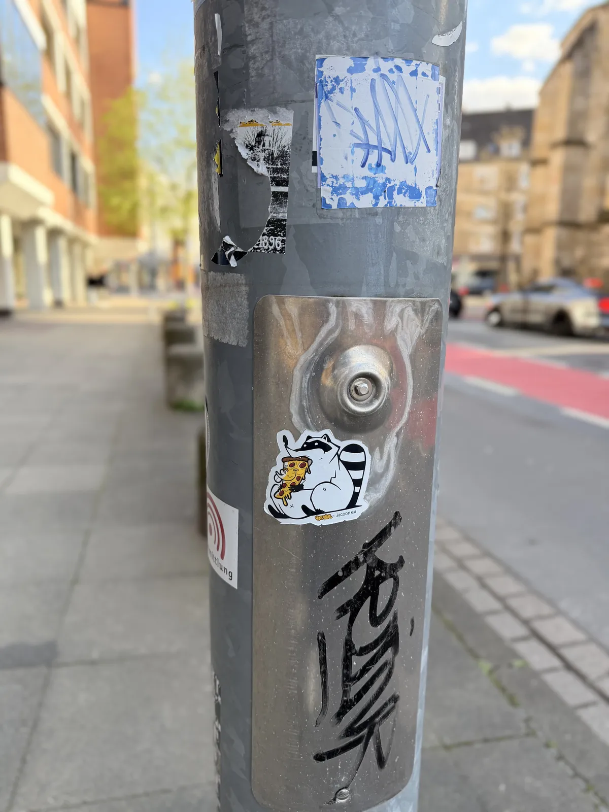 A street photo featuring a gray utility pole that is covered with various stickers, including a cartoon raccoon and some tags. The urban environment in the background consists of buildings and a sidewalk.