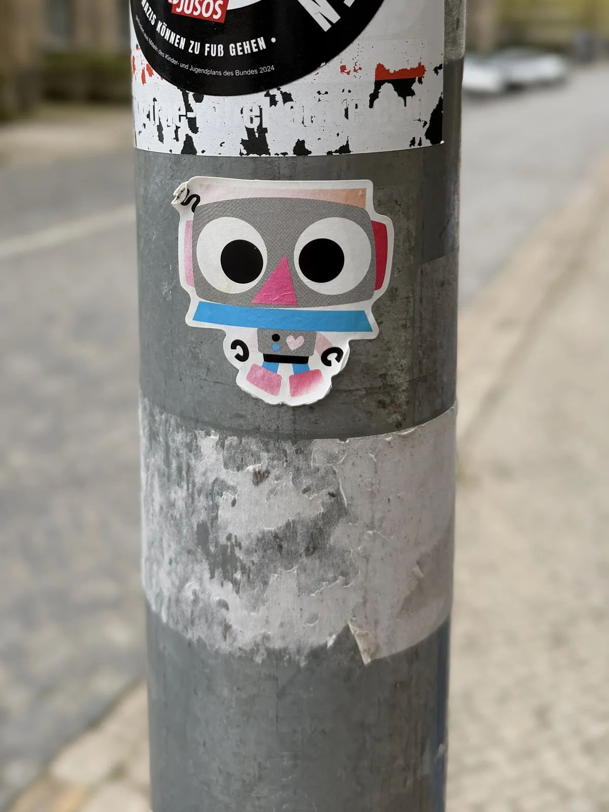 A colorful sticker shaped like a cartoon robot is affixed to a gray utility pole. The robot has large eyes, a triangular nose, and a playful expression, surrounded by layers of other stickers and peeling paper.