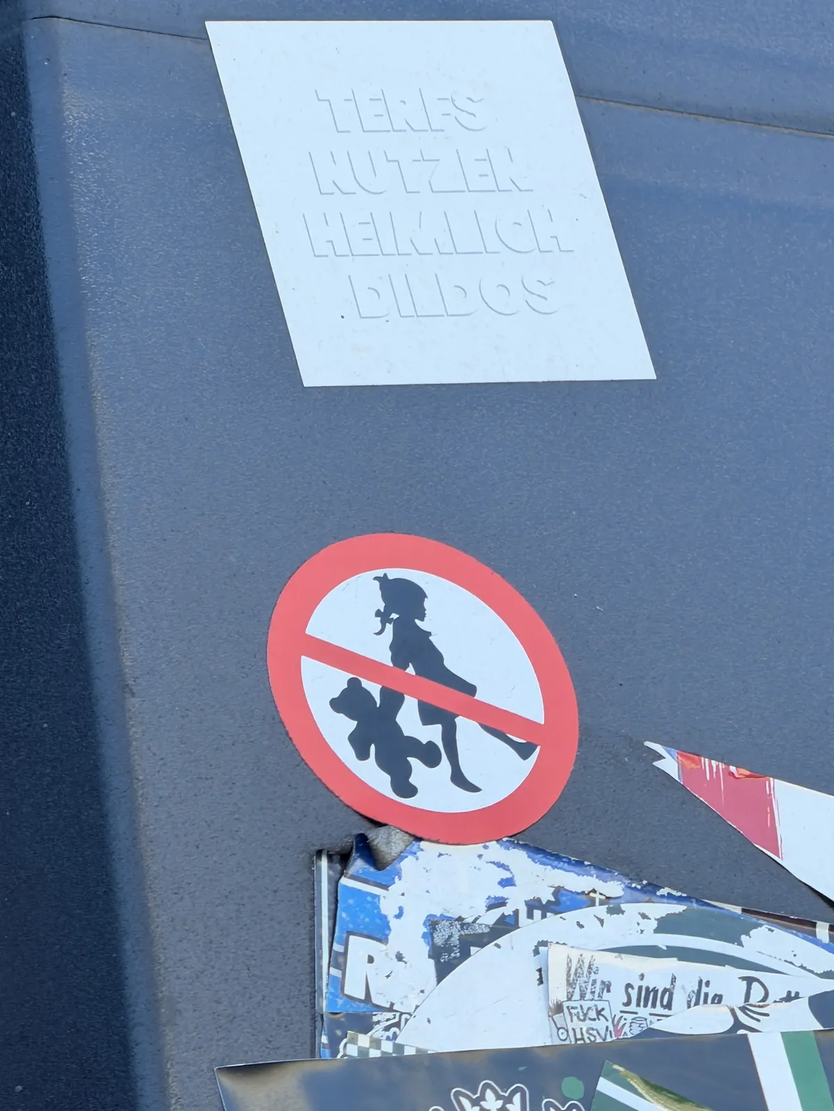 A gray surface covered with various stickers, including a large circular red sticker with a black silhouette of a child holding a teddy bear. There is also a smaller, white sticker with embossed text that is partially readable.