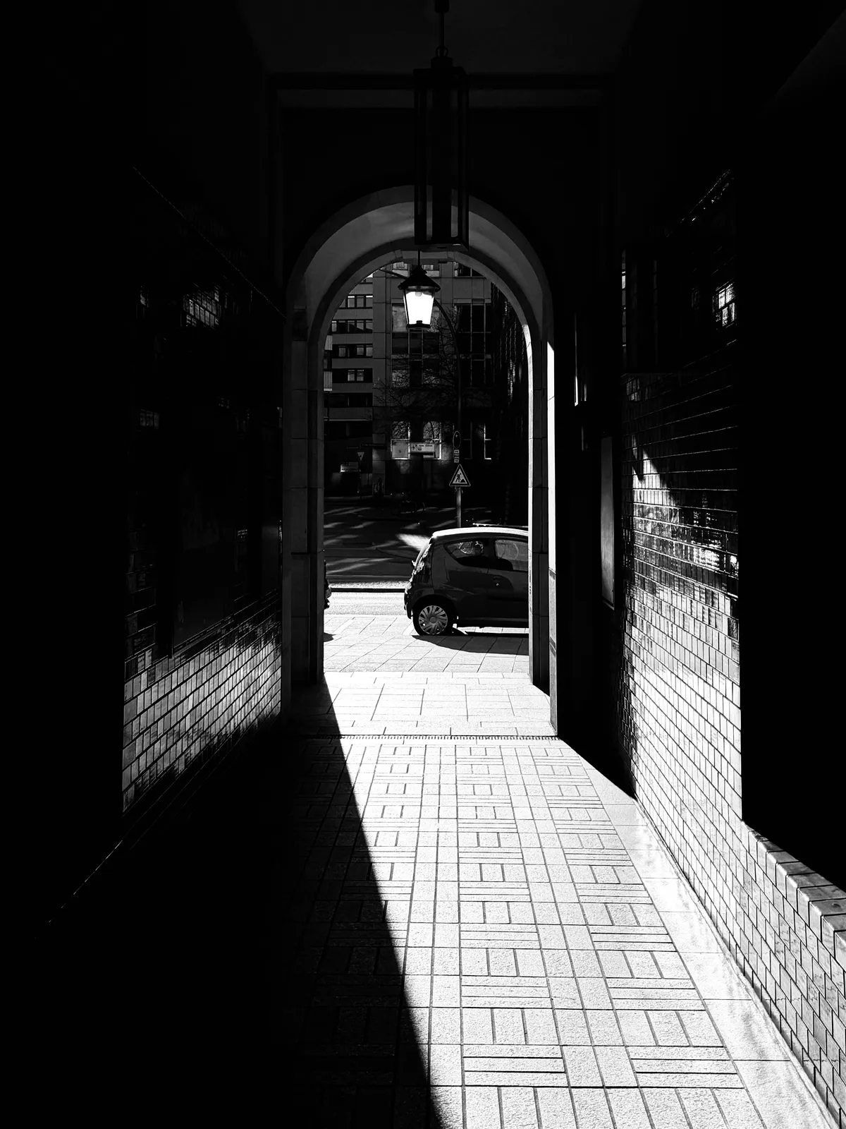 A dark archway leads to a brightly lit street, with a shadow stretching across the tiled floor. A parked car is visible in the distance, illuminated by sunlight.