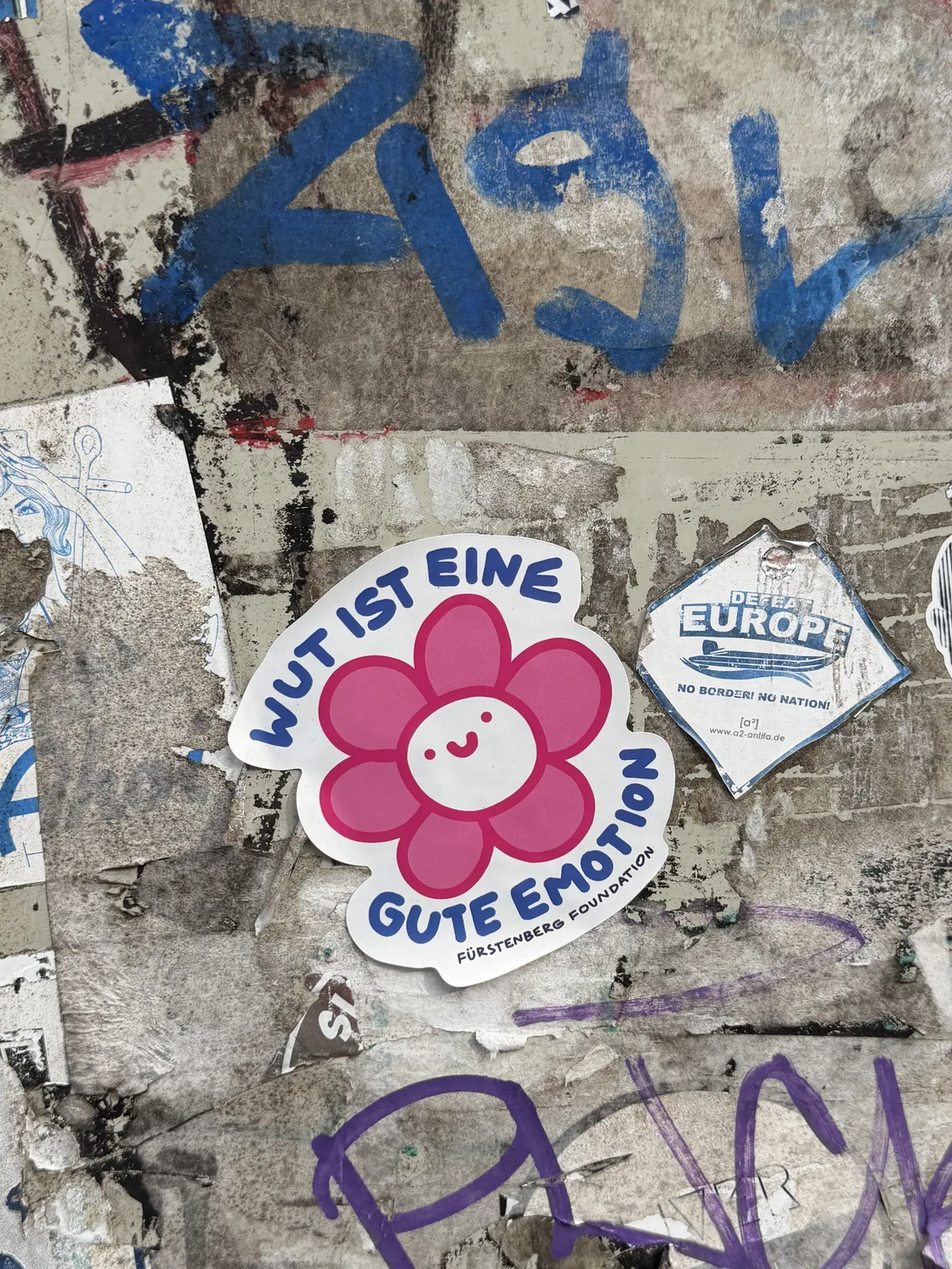 A colorful sticker featuring a smiling pink flower with a round face and the german text 