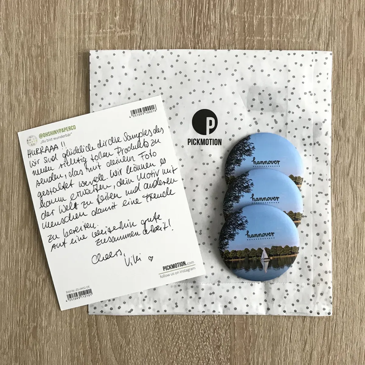 A flat lay of a decorative envelope with a white background covered in small polka dots, accompanied by a postcard featuring handwritten text. Next to the envelope are three round buttons with images of a scenic view, likely depicting a landscape associated with "Hannover."