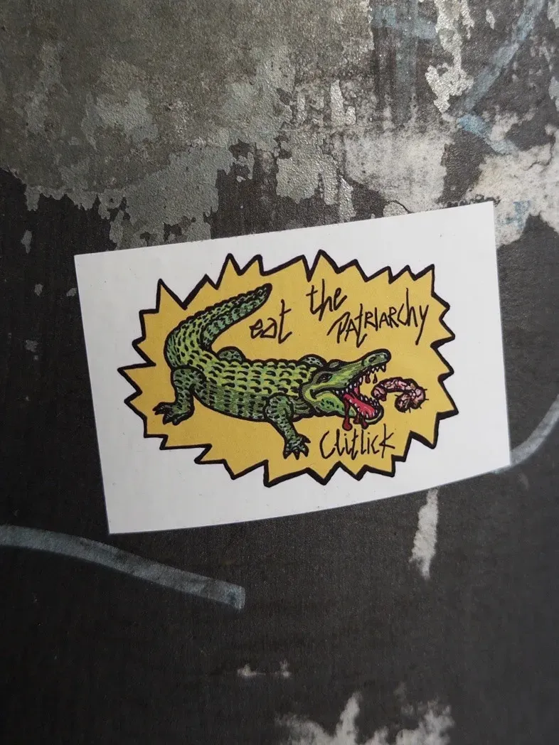 A sticker features a cartoon alligator with its mouth open, holding what appears to be a piece of meat, set against a yellow, jagged background. The text reads 