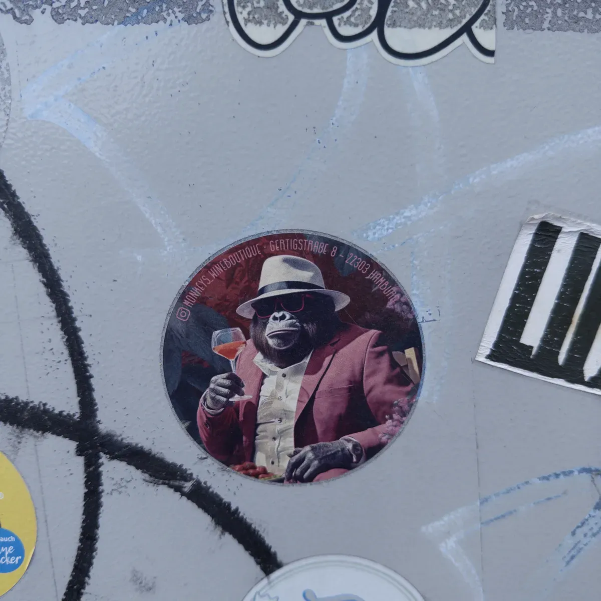 A circular sticker featuring a gorilla dressed in a pink suit and a white hat, holding a cocktail glass. The background is a mix of colorful graffiti and urban textures.