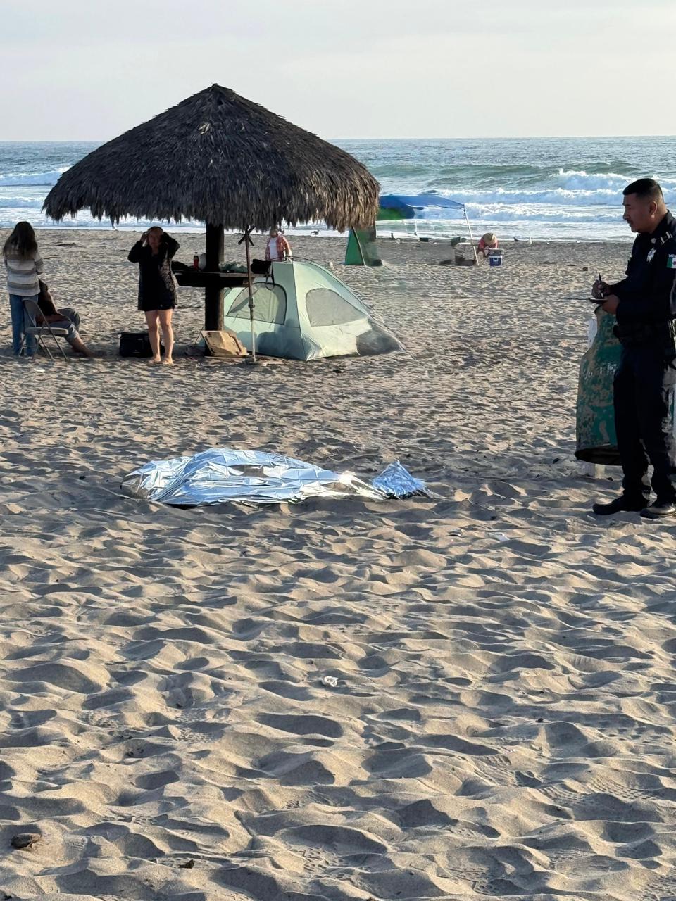 en la imagen se aprecia tendido en la arena el cuerpo del hombre que murió ahogado en la playa La Misión, cubierto con una manta, y un  oficial de policía de pie junto al cuerpo. Al fondo se observa el mar, casas de campaña y algunas personas caminando en la arena