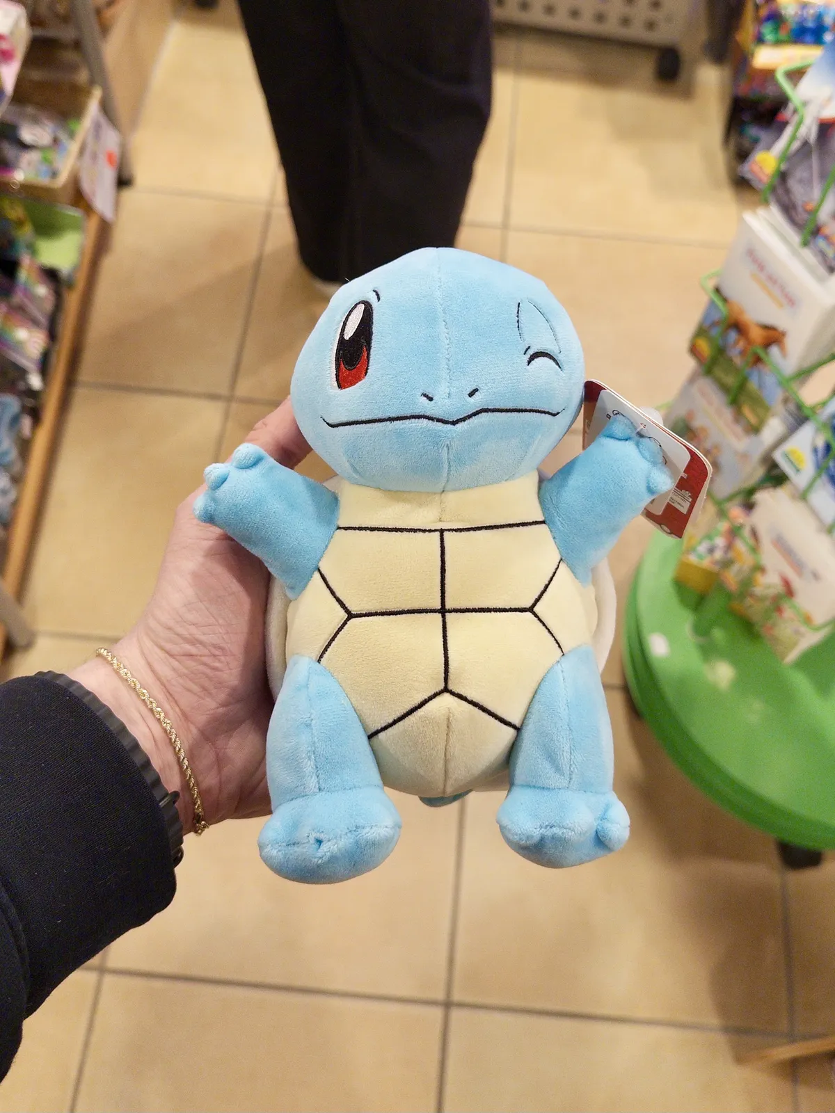 A person holding a light blue plush toy of a turtle-like character with large eyes and a friendly expression, featuring a shell with yellow and black markings. The background shows shelves stocked with various toys and merchandise in a store.