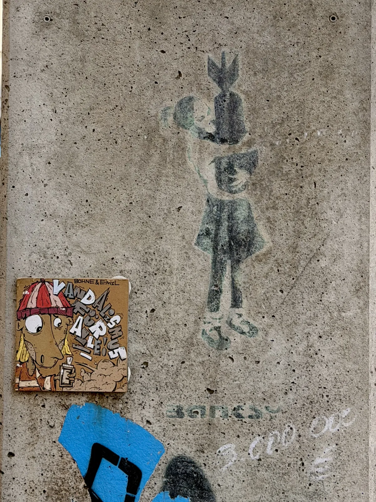 A faded graffiti by Banksy depicting a girl holding a bomb, aka "The Bomb Hugger". The wall is textured concrete with graffiti and markings.