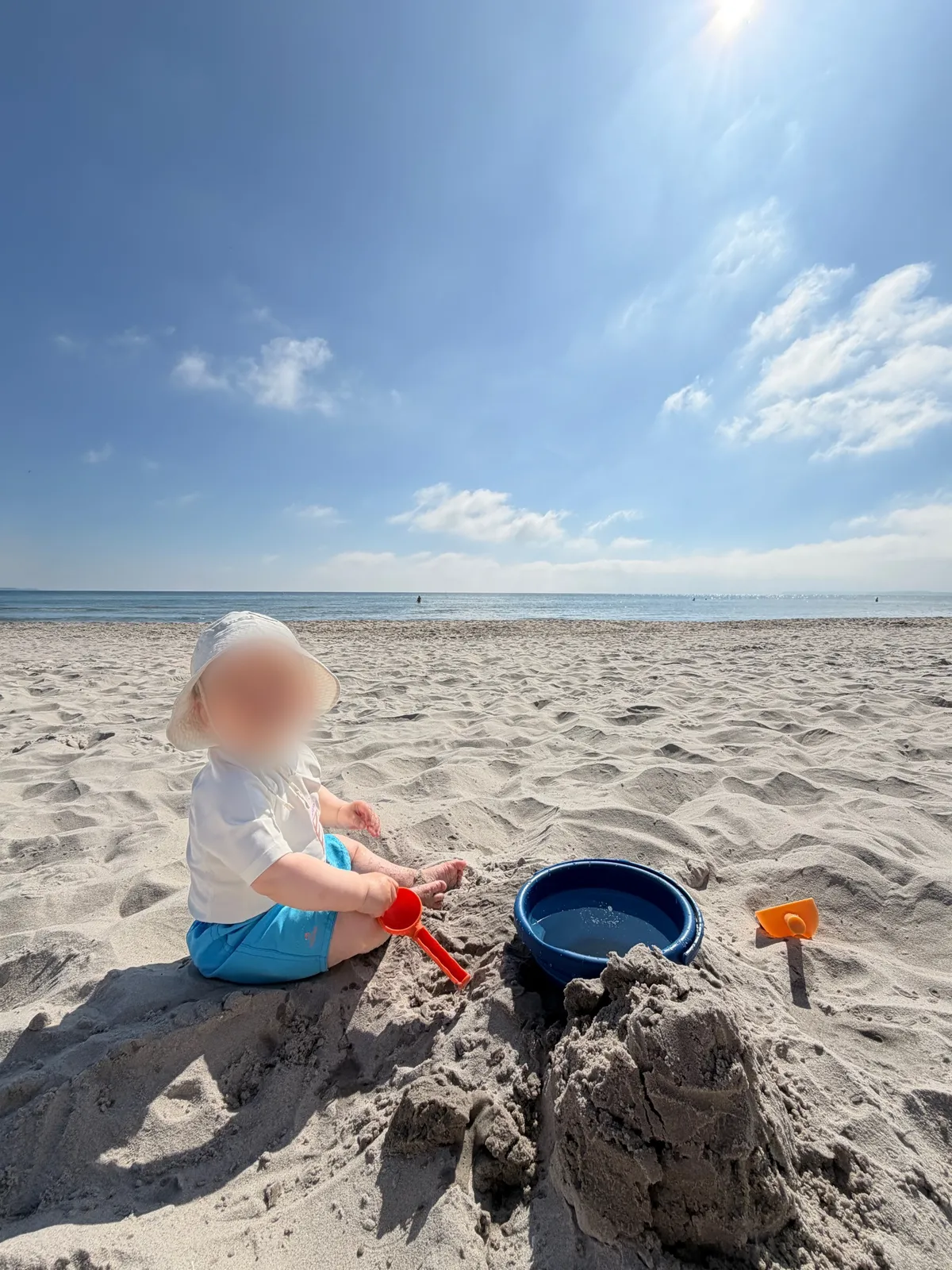 A child wearing a sun hat sits on a sandy beach, playing with a blue bucket and a red shovel. The ocean stretches out in the background under a clear blue sky.