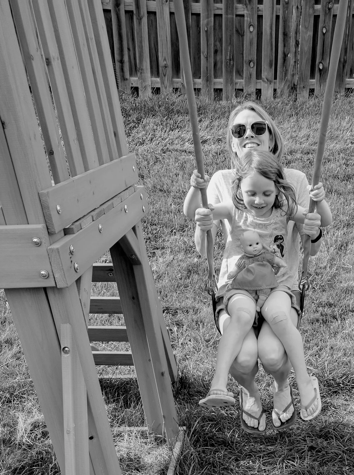 This black-and-white photo captures a sweet moment of joy. My wife is on a backyard swing with my stepdaughter sitting on her lap, holding a doll. They both look like they’re having fun, with my wife smiling broadly while the swing moves. The wooden swing set and grassy yard create a cozy, playful vibe.