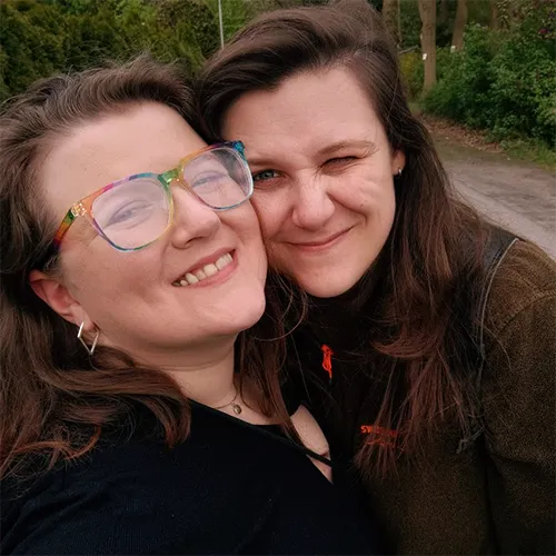 A photo of my wife and I. My wife is on the left, with brown hair and rainbow glasses on, wearing a toothy grin. She has a black top on and silver earrings that resemble a diamond shape. I am on the right, with a similar brown hair colour, winking with my left eye and also smiling, although with my mouth closed. I am wearing a brown jacket, small silver hoop earrings and you can see the straps of my black backpack. In the background you can see some trees and green bushes.